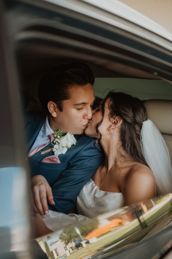 A man wearing a blue suit with white flowers on his lapel leans over to kiss a woman who is wearing a white dress. They are sitting in the back of a car together. The background shows an outdoor scene with trees and grass.