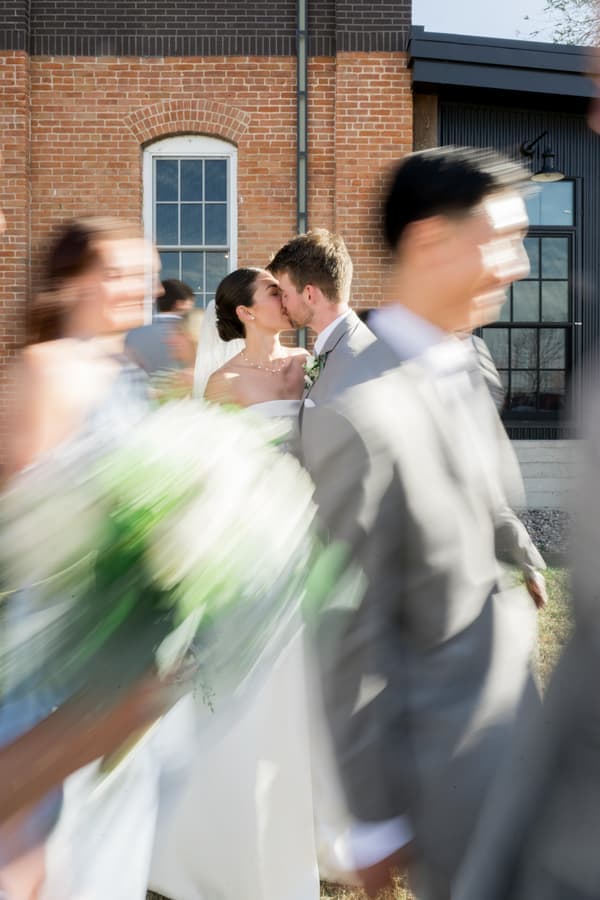 A bride and groom share a kiss amidst blurred figures at an outdoor wedding ceremony.