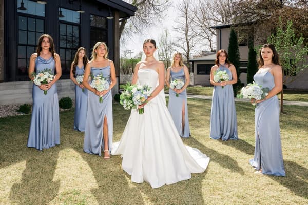 a bride and her bridesmaids in formal attire pose for a group photo outdoors on a sunny day.