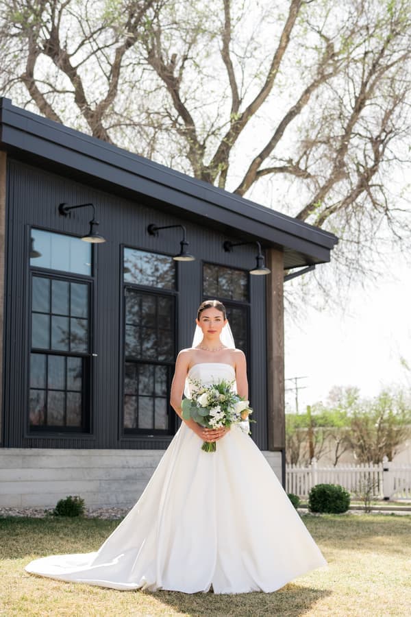 a bride in a white wedding dress stands outdoors holding a bouquet, with a modern black building and trees in the background.